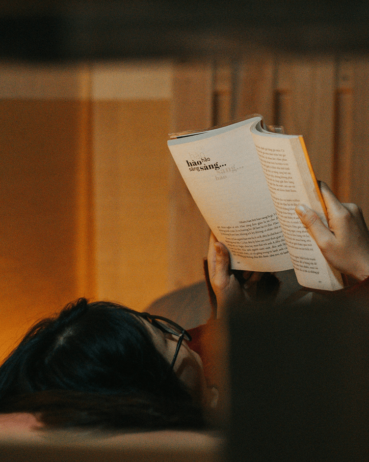 A person lying in bed at night, holding a book above them and reading by warm ambient light.